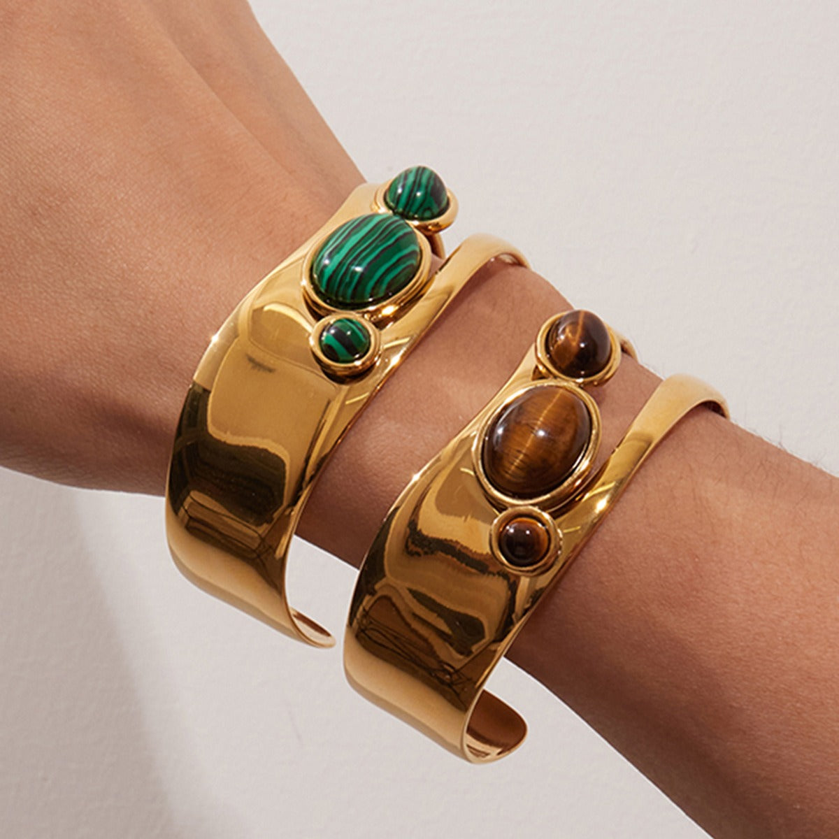 A model's wrist adorned with gold cuff bracelets featuring malachite and tiger's eye stones, demonstrating their stylish and stackable design.