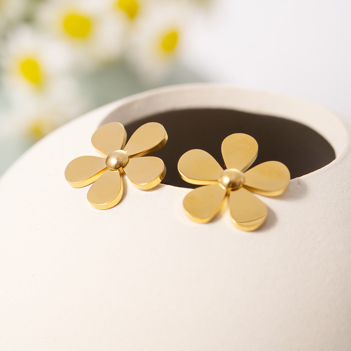 A pair of gold daisy flower stud earrings resting on a white display holder.