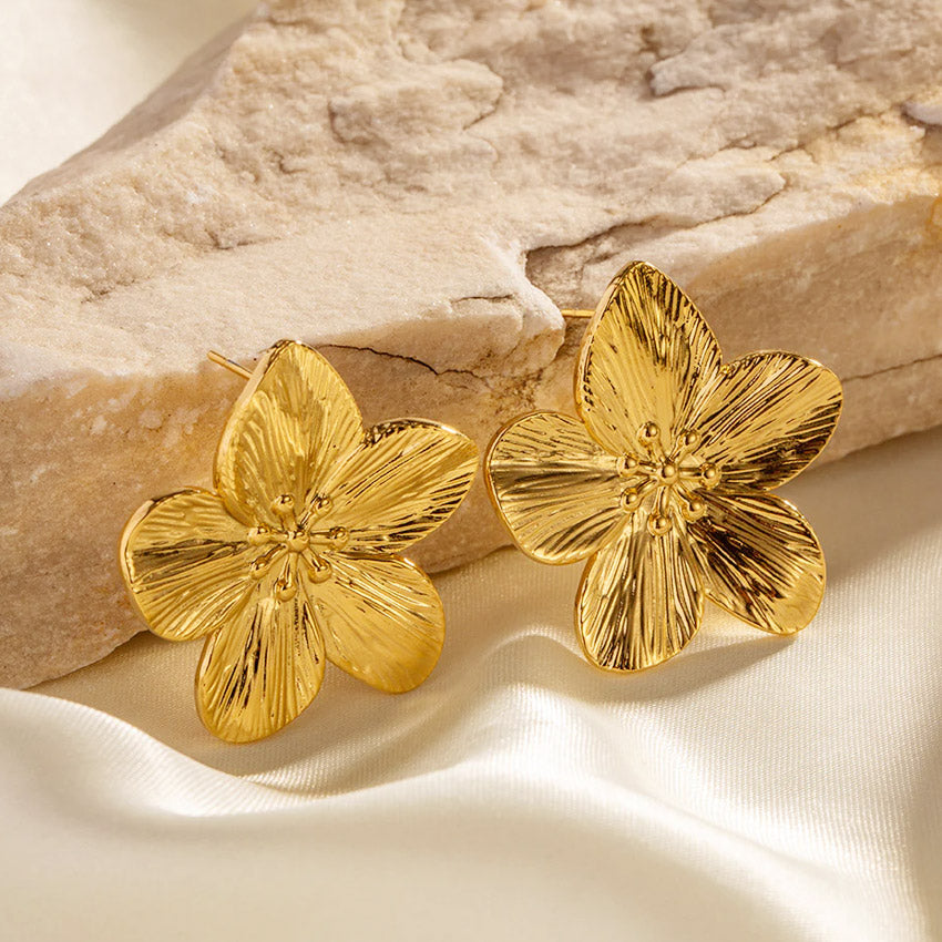 A pair of gold blossom flower earrings with a textured surface displayed on a neutral fabric background.
