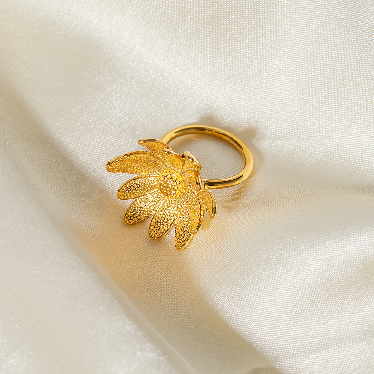A close-up of the gold-finished stainless steel daisy ring on a white fabric background.