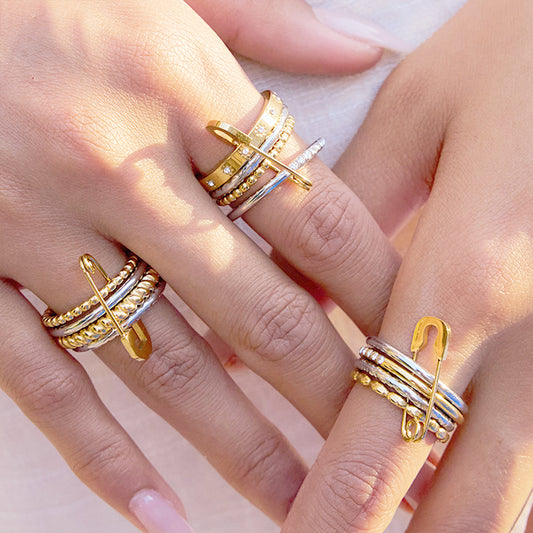 A variety of stackable gold and silver tone stainless steel rings with safety pin charm on a model's hand.