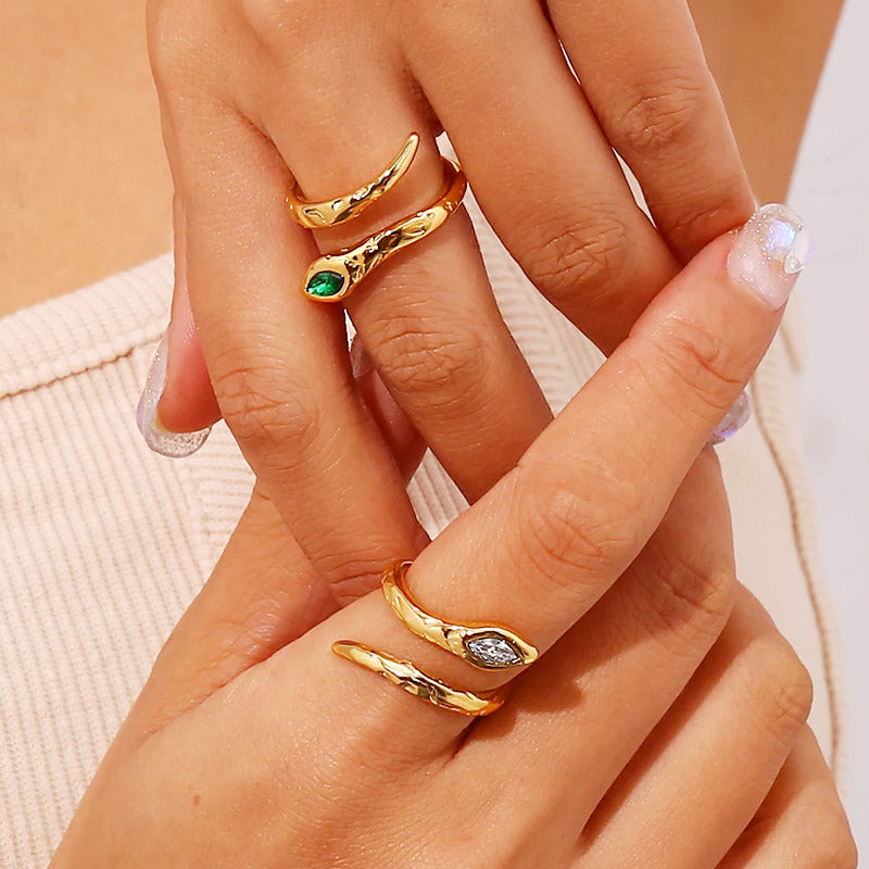 A model's hand showcasing multiple gold snake rings with green and clear gemstones.