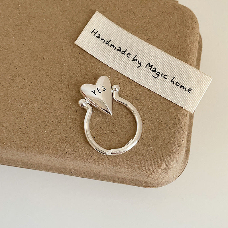 A silver adjustable ring with a YES heart design on a finger