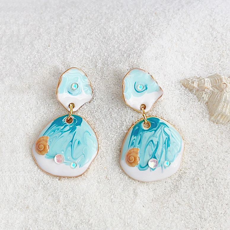 A detailed view of the ocean-themed earrings with marbled blue enamel and golden seashell accents.