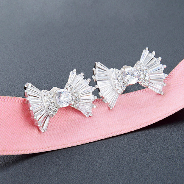 A pair of silver bow earrings with clear gemstones presented on a delicate pink ribbon
