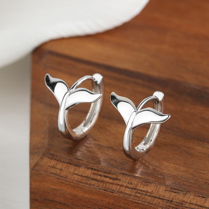 A pair of silver mermaid fishtail huggie hoop earrings displayed on a wooden surface.