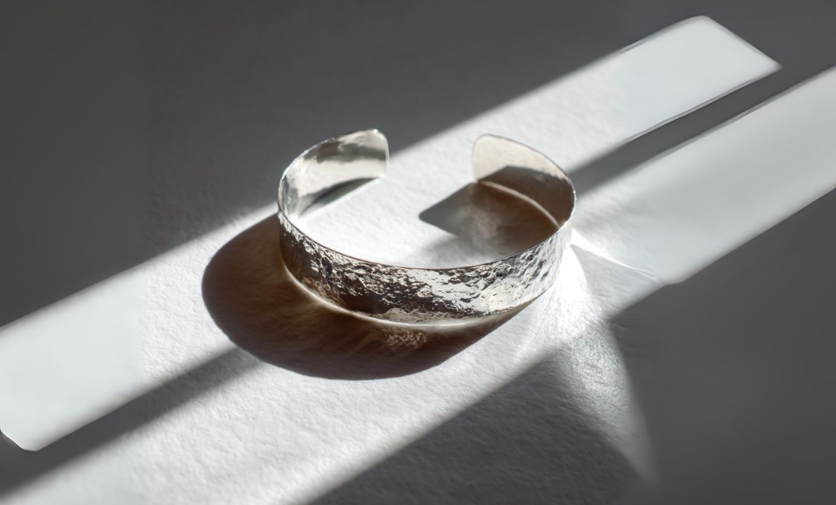 Polished silver bracelet with hammered texture reflecting natural light on white background.